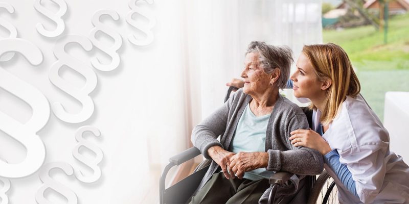 Eine Seniorin im Rollstuhl schaut mit ihrer Pflegekraft aus dem Fenster; links daneben Paragraphensymbole