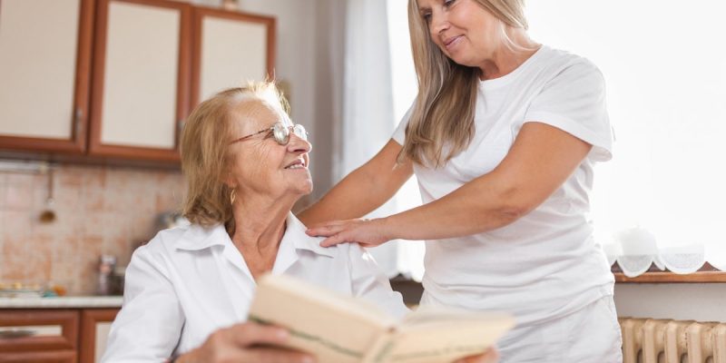 Eine Seniorin sitzt mit ihrem Buch am Küchentisch und wird von einer Pflegekraft umsorgt